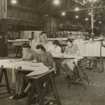 Workers in a textile cutting room laying long bolts of cotton fabric on a stainless steel table beneath even daylight, with garment patterns pinned and bundled at the side.